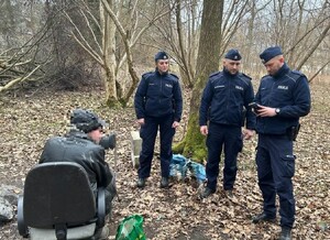 Trzech funkcjonariuszy Szkoły Policji, dwóch policjantów i policjantka, w umundurowaniu dokonują legitymowania mężczyzny siedzącego na krześle. Teren leśny
