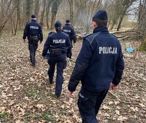 Trzech funkcjonariuszy Szkoły Policji, dwóch policjantów i policjantka, w umundurowaniu dokonują legitymowania mężczyzny siedzącego na krześle. Teren leśny